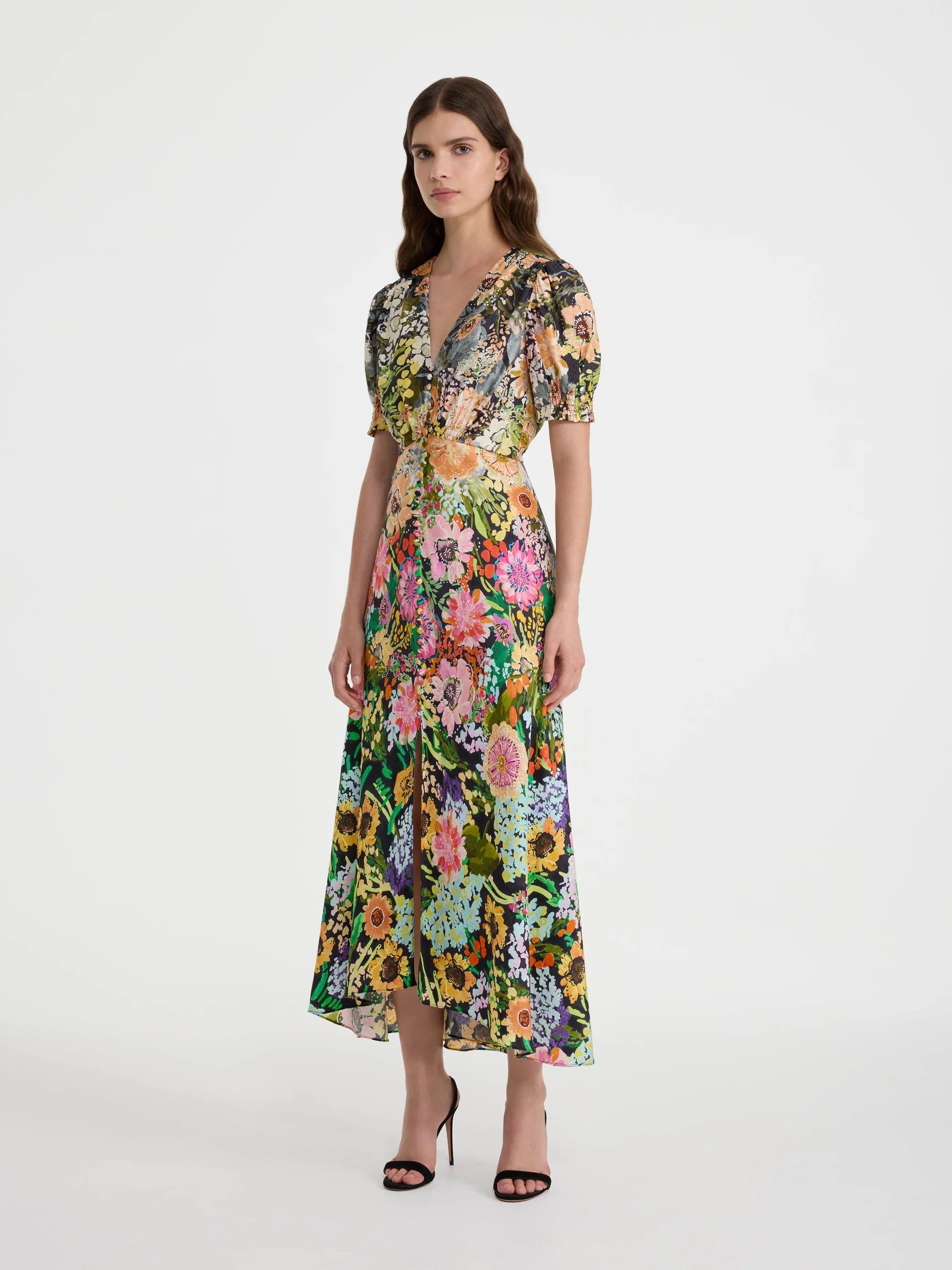 A model wearing a short-sleeved midi dress with a vibrant multicolor floral print, standing against a plain light gray studio background.