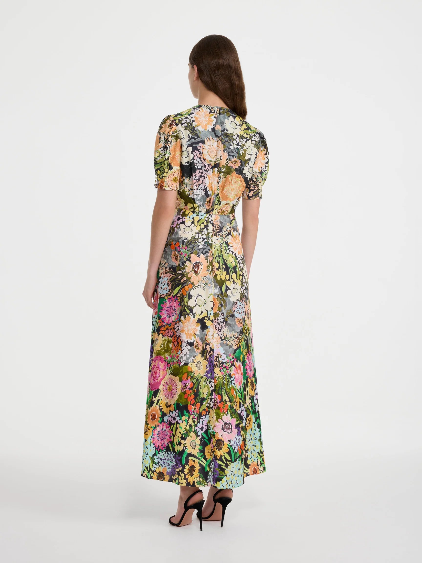 A woman viewed from the back wearing a short-sleeve maxi dress with a dense multicolor floral print on a dark background, paired with black high heels, against a plain light gray studio backdrop.