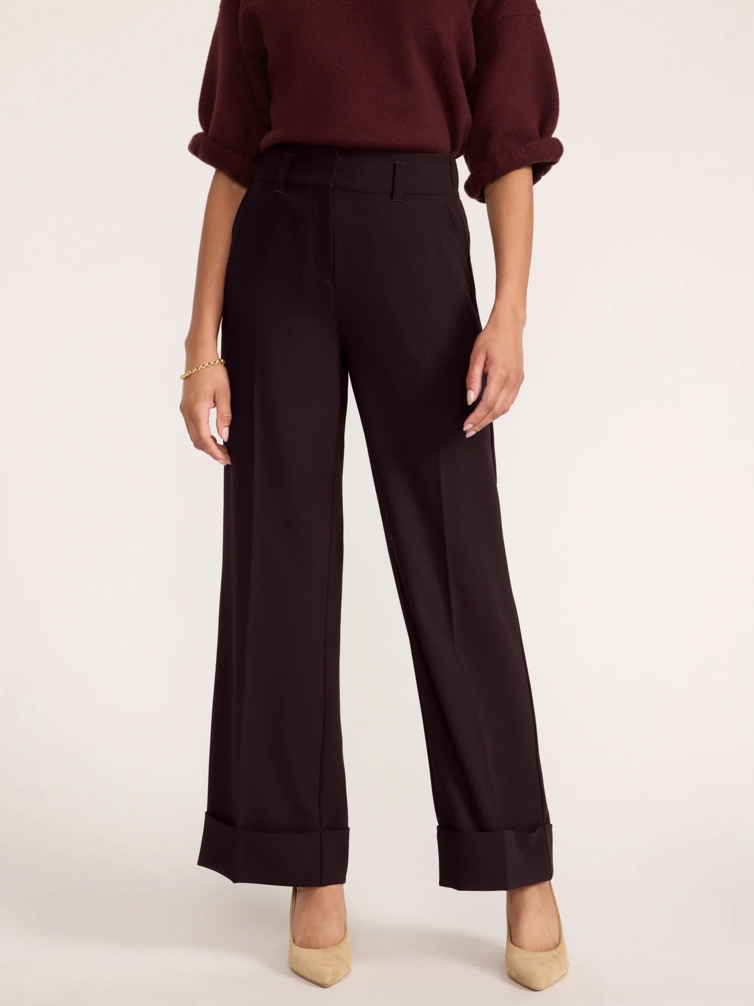 A model is shown from the chest down wearing dark brown wide-leg trousers with rolled cuffs, paired with a dark burgundy top and beige heels, against a plain off-white studio background.