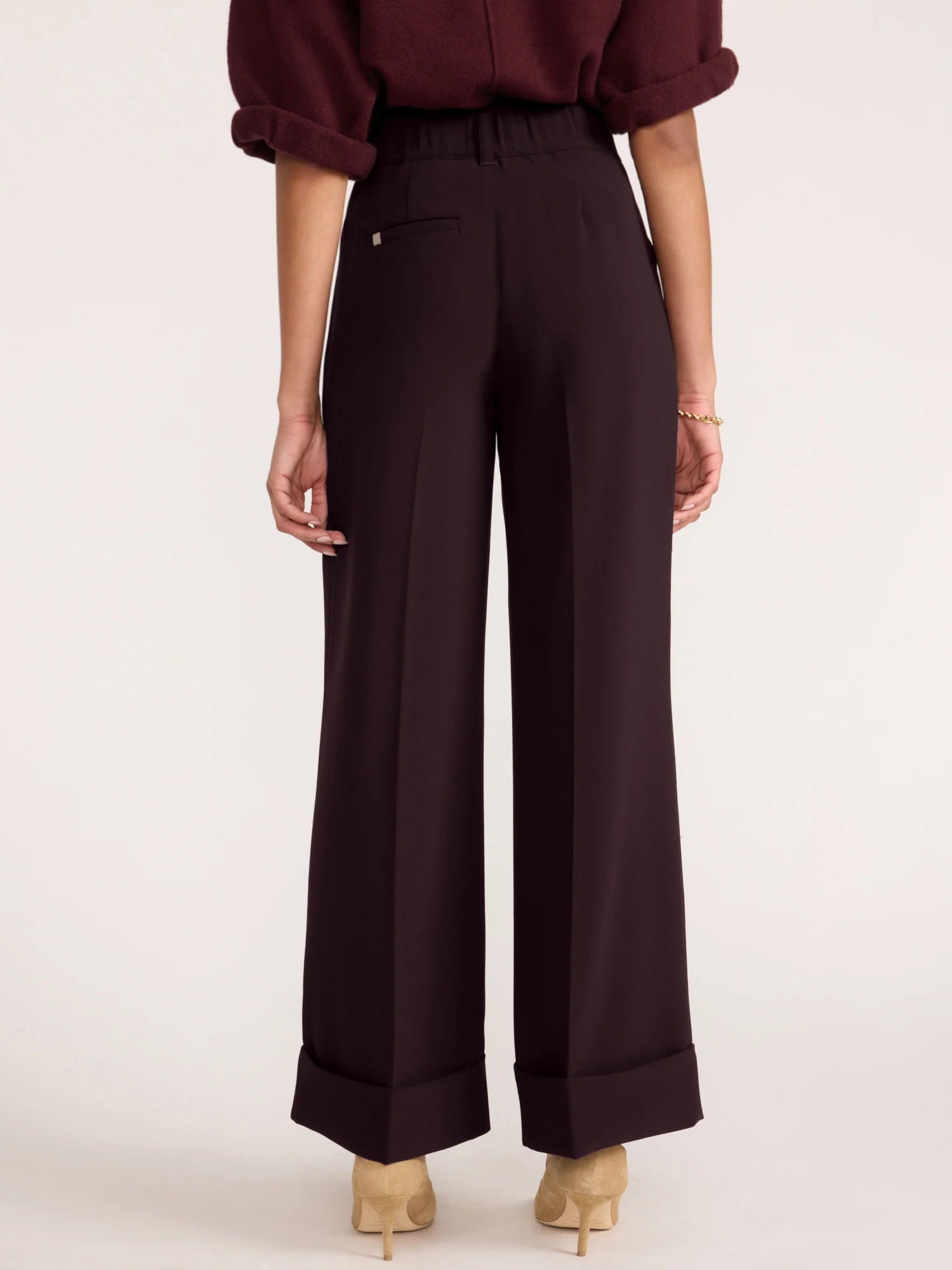 A model shown from the back wearing dark plum wide-leg trousers with rolled cuffs, paired with a matching dark plum blouse and beige heels, against a plain off-white studio background.