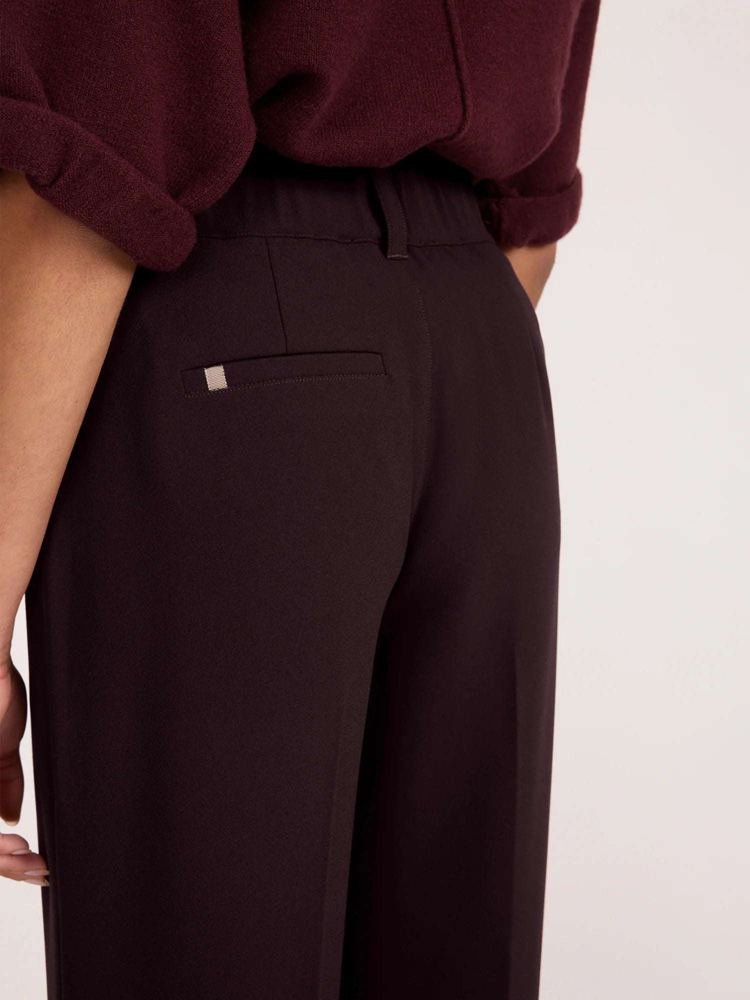 Close-up back view of a person wearing dark plum wide-leg trousers with a small welt pocket and a dark burgundy top, against a plain light background.