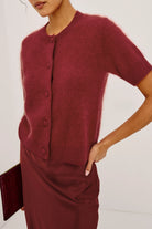 A close-up of a model wearing a short-sleeve fuzzy knit cardigan and matching satin skirt in a deep burgundy red, holding a matching clutch.