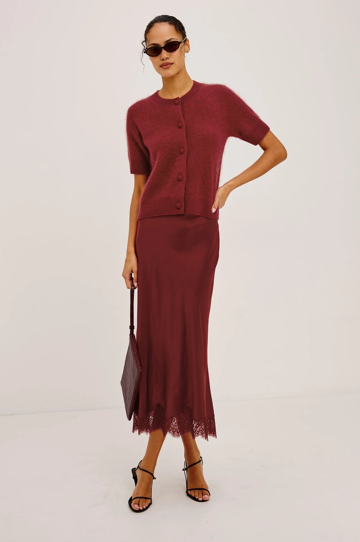A model wearing a matching burgundy knit short-sleeve cardigan and midi skirt set, styled with sunglasses, a dark handbag, and black strappy heels against a plain white studio background.