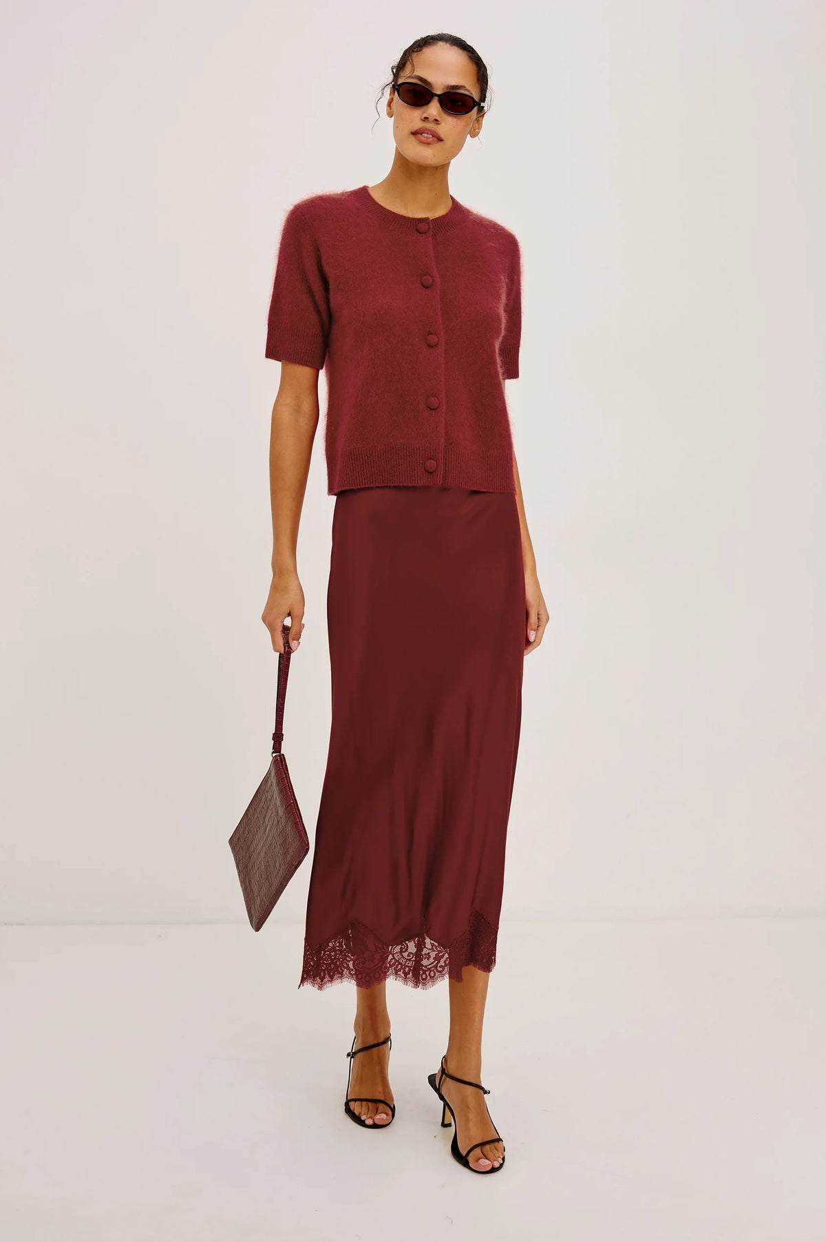 A model stands against a plain white studio background wearing a matching burgundy two-piece set consisting of a short-sleeve cropped button-front cardigan and a long midi skirt with lace trim. She is also wearing black strappy heels and holding a matching burgundy handbag.