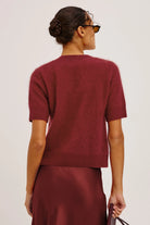 A woman shown from the back wearing a short-sleeve knit sweater in a deep burgundy red, paired with a matching satin skirt, against a plain light gray studio background.