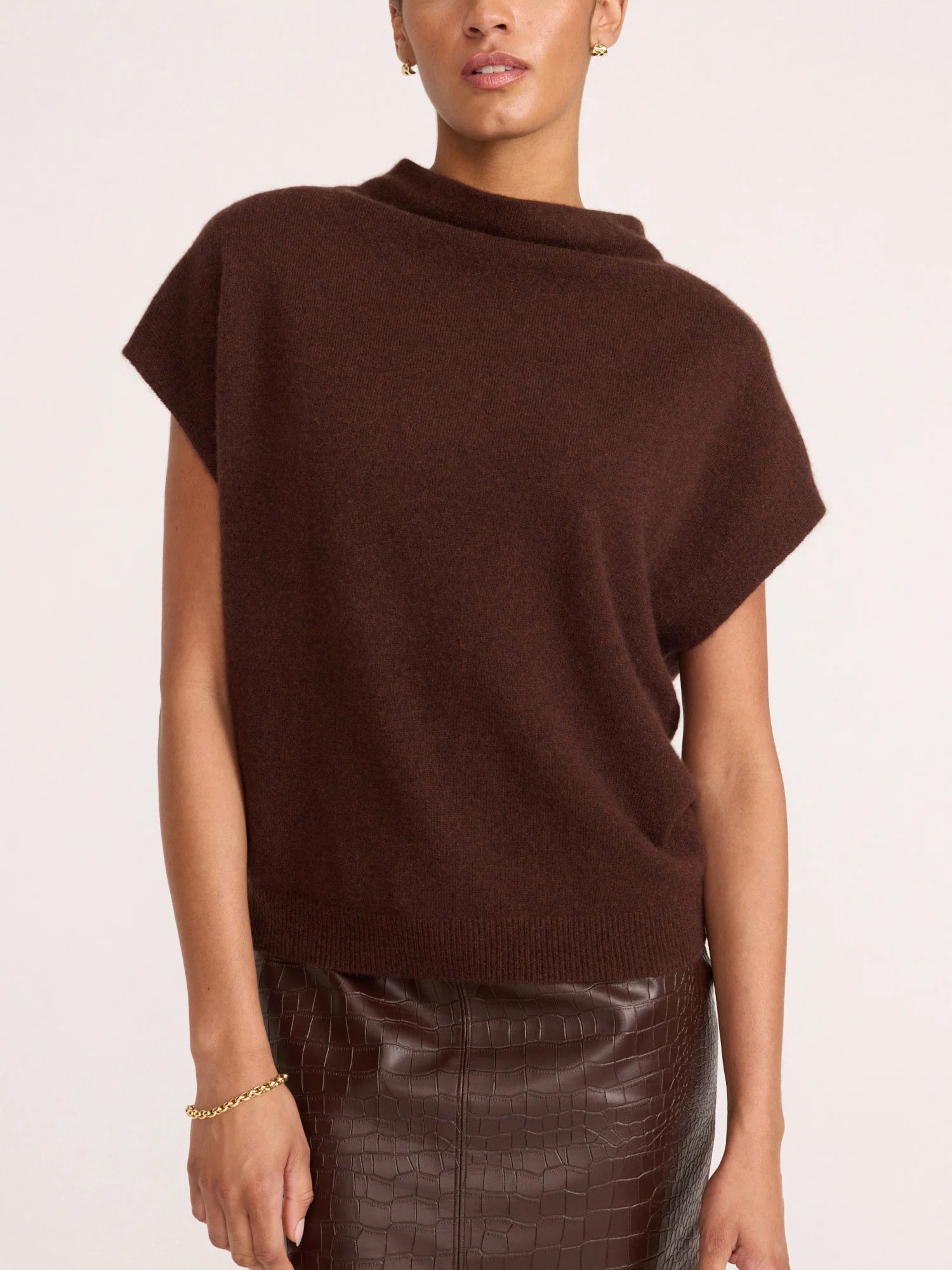 A woman wearing a dark brown knit sweater with a wide draped neckline and short dolman sleeves, paired with a dark brown textured skirt, against a plain light background.