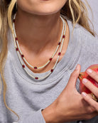 Close-up of a person wearing a layered necklace with red and clear stones, holding a red apple.