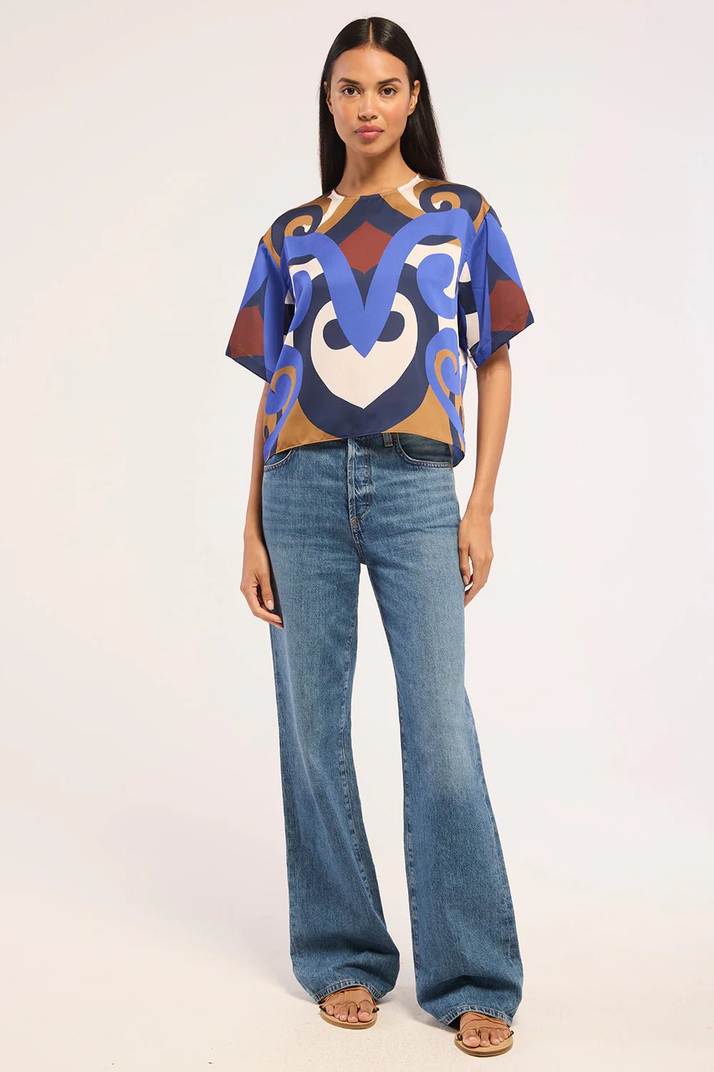 A woman modeling a short-sleeve crop top with a bold abstract pattern in blue, brown, beige, and white, paired with wide-leg blue jeans and tan sandals, against a plain light background.