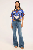 A woman modeling a short-sleeve crop top with a bold abstract pattern in blue, brown, beige, and white, paired with wide-leg blue jeans and tan sandals, against a plain light background.