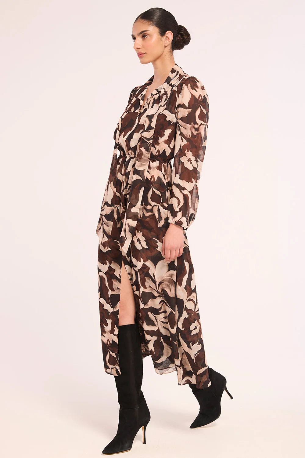 A woman modeling a long-sleeved midi dress with a brown, beige, and white abstract floral pattern, paired with black knee-high heeled boots, against a plain light background.