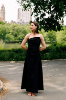 A woman wearing a strapless black floor-length gown stands outdoors on a paved path in a park, with trees, greenery, and buildings in the background.