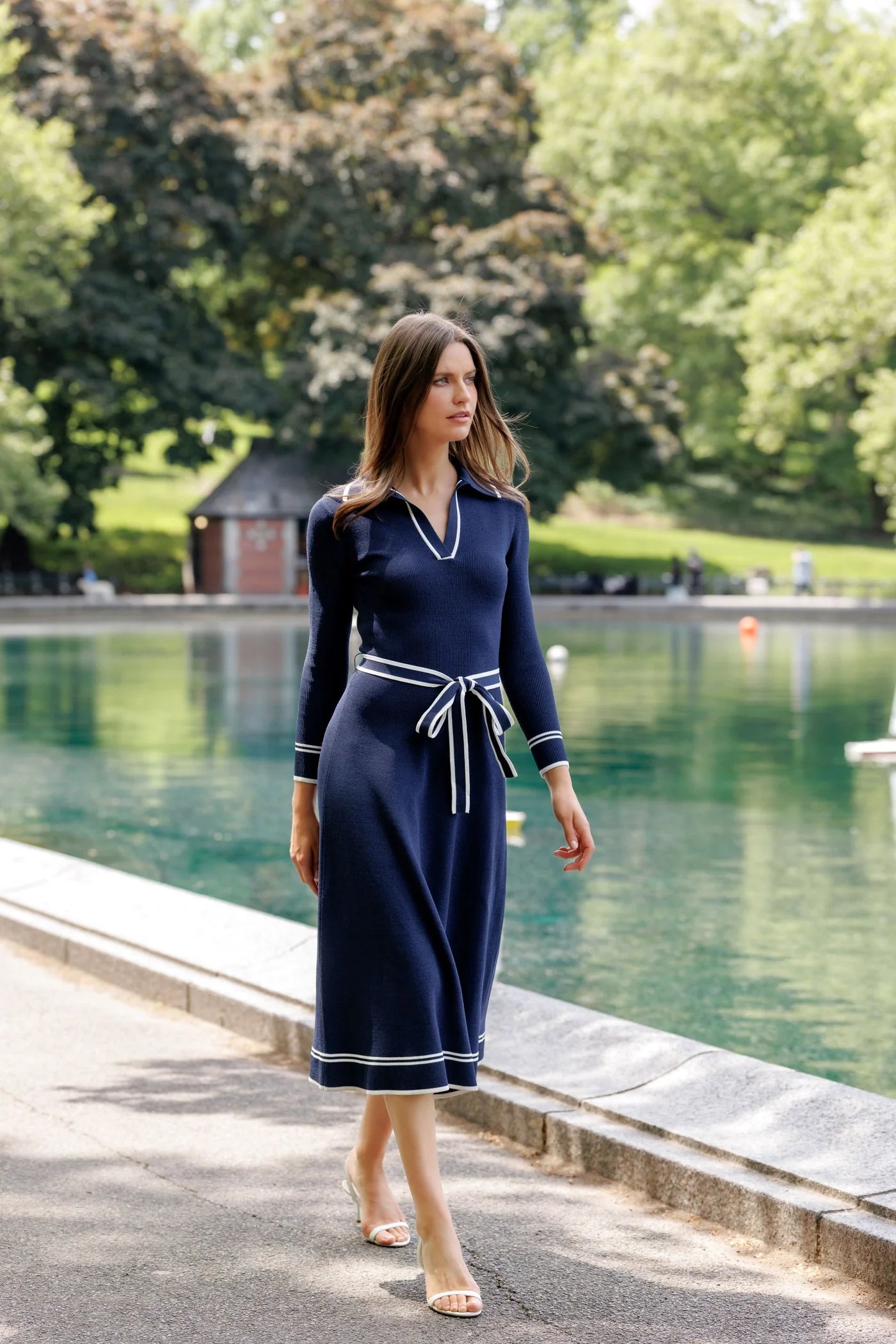 A woman wearing a navy blue midi dress with white piping details walks along a paved path next to a green park pond.