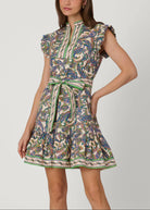 A woman modeling a sleeveless mini dress with a high mandarin collar, ruffled cap sleeves, a self-tie waist belt, and a colorful paisley pattern on a light beige base.