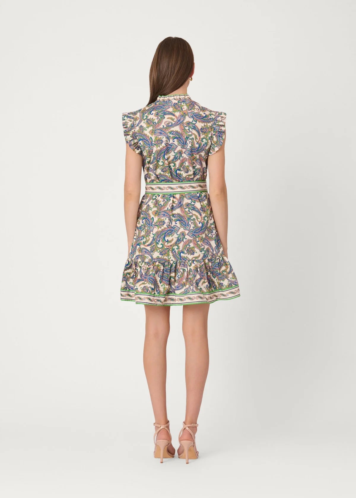 A woman viewed from the back wearing a short ruffled dress with a multicolor paisley pattern in blue, green, yellow, and beige tones, paired with beige strappy heels, against a light gray studio background.
