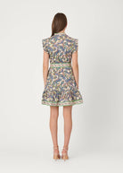 A woman viewed from the back wearing a short ruffled dress with a multicolor paisley pattern in blue, green, yellow, and beige tones, paired with beige strappy heels, against a light gray studio background.