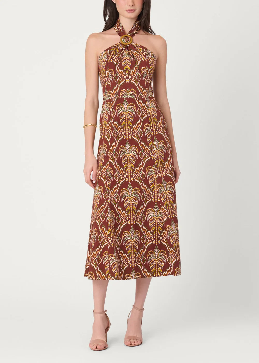 A woman modeling a halter-neck midi dress with a brown, gold, and beige palm tree pattern against a plain light gray studio background.