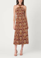 A woman modeling a halter-neck midi dress with a brown, gold, and beige palm tree pattern against a plain light gray studio background.