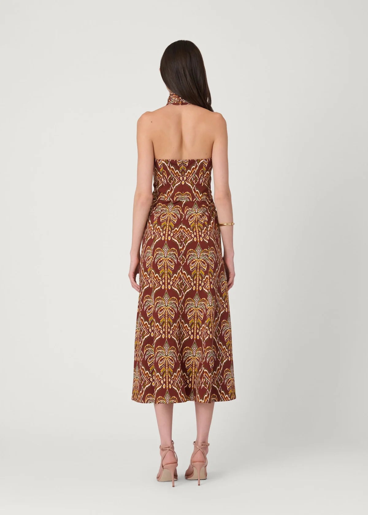 A woman viewed from the back wearing a strapless midi dress with a brown, gold, and beige palm tree pattern, paired with nude high-heeled sandals against a light gray studio background.