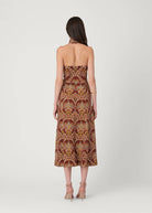 A woman viewed from the back wearing a strapless midi dress with a brown, gold, and beige palm tree pattern, paired with nude high-heeled sandals against a light gray studio background.