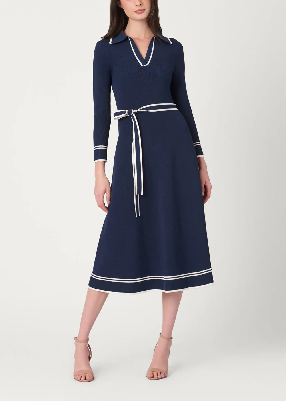 A woman modeling a navy blue midi dress with white contrast trim, a matching waist tie belt, and white piping details on the collar and cuffs, set against a plain light gray studio background.
