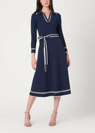 A woman modeling a navy blue midi dress with white contrast trim, a matching waist tie belt, and white piping details on the collar and cuffs, set against a plain light gray studio background.
