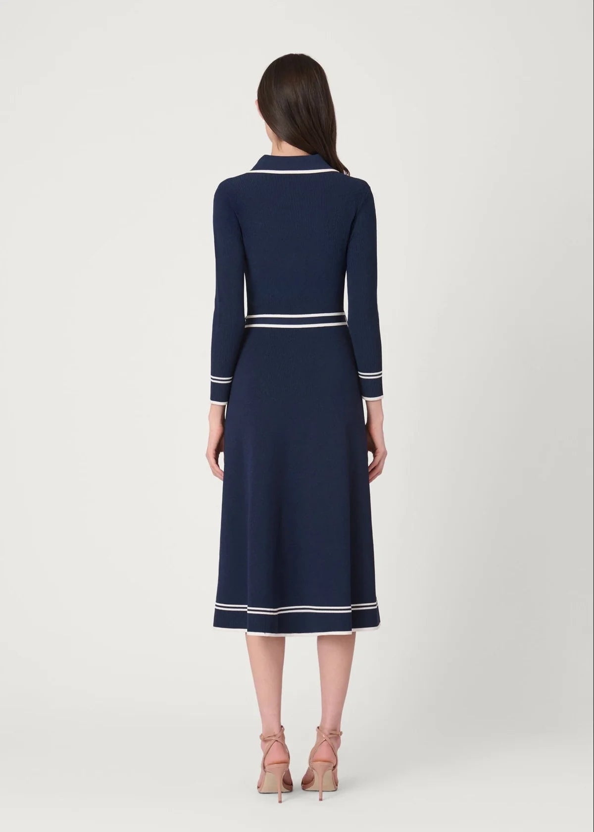 A woman viewed from the back wearing a navy midi dress with white contrast trim at the collar, cuffs, and hem, paired with nude high-heeled sandals against a light gray studio background.