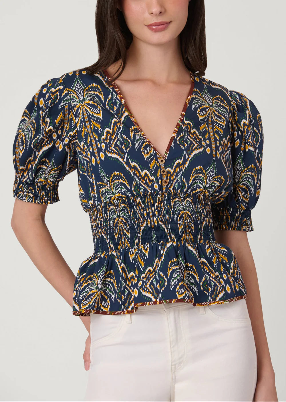 A woman wearing a navy blue short-sleeve blouse with a yellow and gold abstract pattern, paired with white pants, against a light gray studio background.