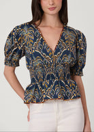 A woman wearing a navy blue short-sleeve blouse with a yellow and gold abstract pattern, paired with white pants, against a light gray studio background.