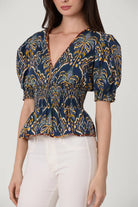 Woman wearing a patterned blue and yellow top with white pants on a plain background