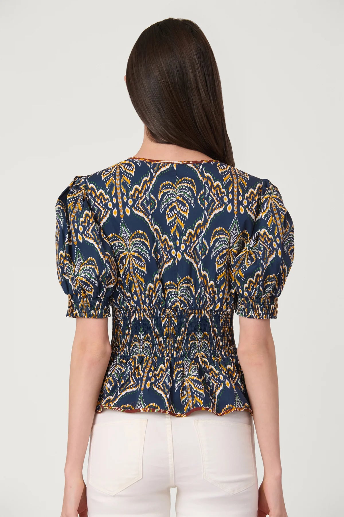 A woman viewed from the back wearing a navy blue short-sleeve blouse with a gold and yellow palm tree pattern and puff sleeves, paired with white pants, against a light gray background.