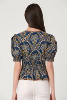 A woman viewed from the back wearing a navy blue short-sleeve blouse with a gold and yellow palm tree pattern and puff sleeves, paired with white pants, against a light gray background.