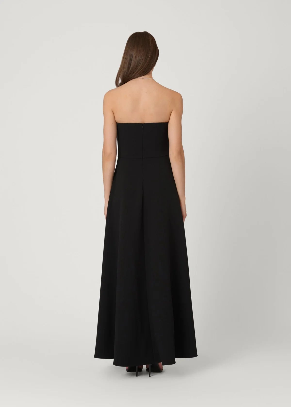 A woman viewed from the back wearing a strapless black maxi dress with a structured bodice and full skirt, standing against a light gray studio background.