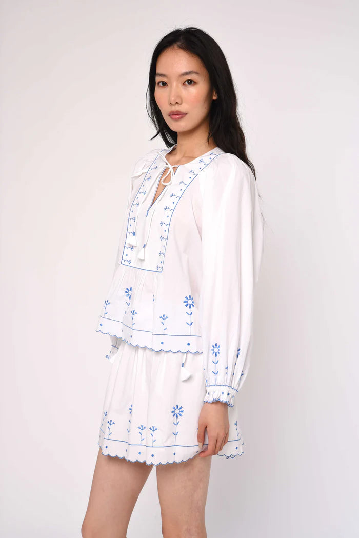 Woman wearing a white blouse with blue floral embroidery on a plain background