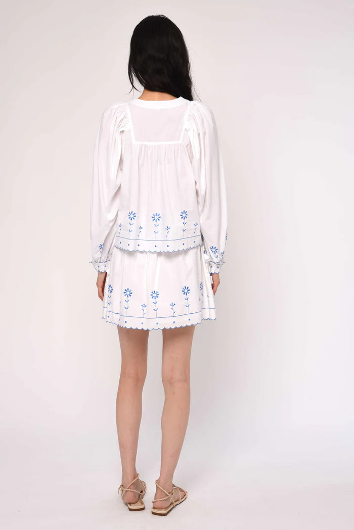 Woman wearing a white outfit with blue floral embroidery on a plain background