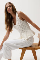 Woman wearing a white crochet top and white pants sitting on a wooden chair against a white background