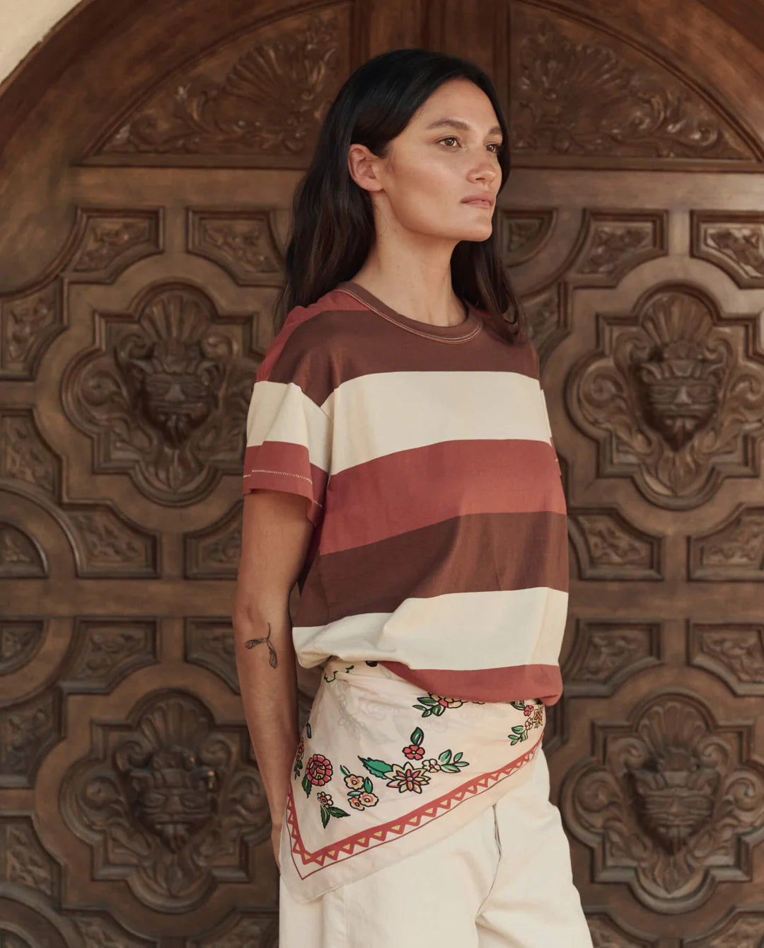 Woman wearing a striped shirt and embroidered skirt standing in front of a wooden door with intricate carvings.