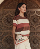 Woman wearing a striped shirt and embroidered skirt standing in front of a wooden door with intricate carvings.