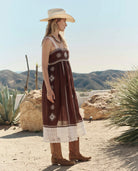 Woman in a brown dress with white patterns and a beige hat standing in a desert landscape.