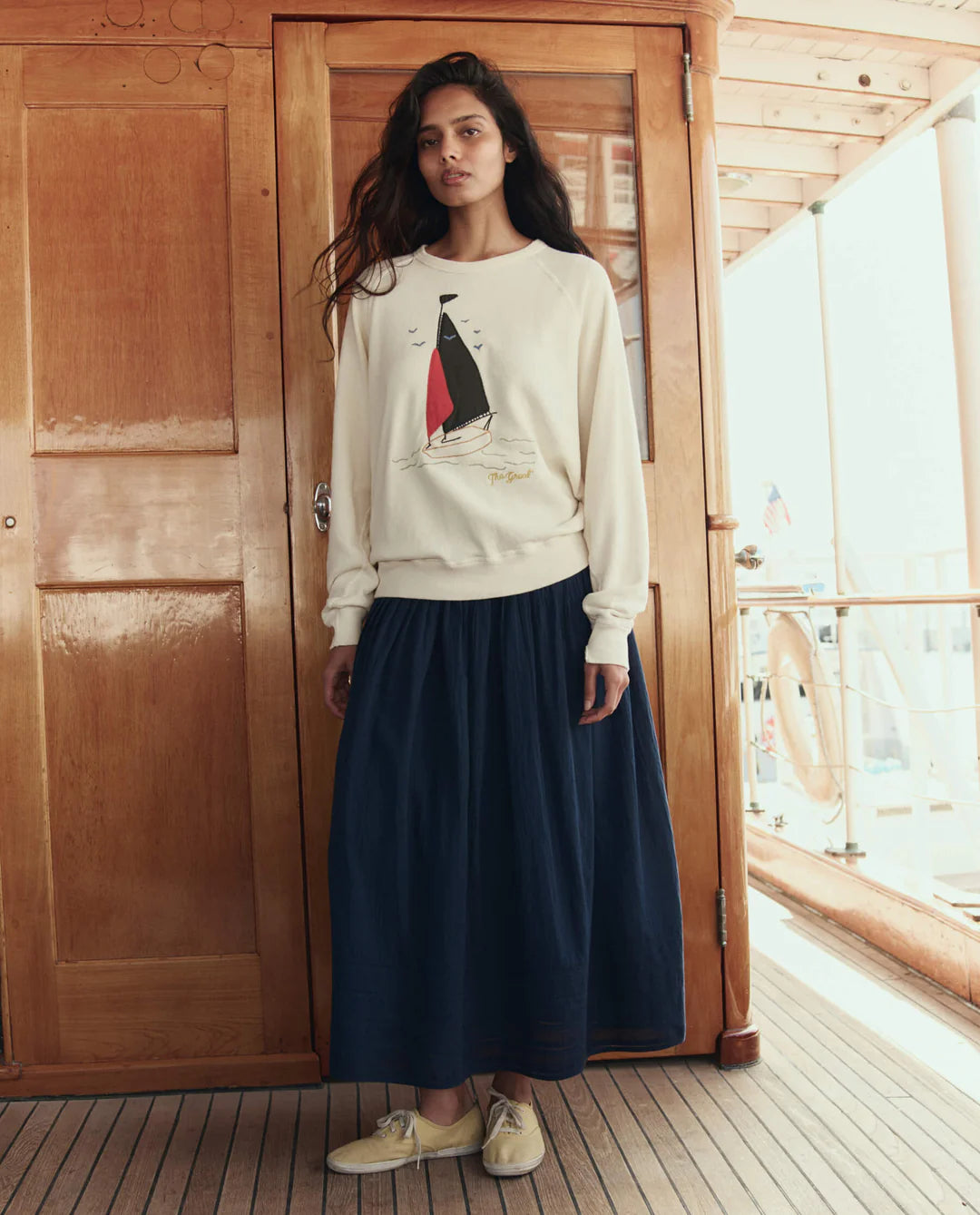 Woman wearing a white sweatshirt with a sailboat design and a navy skirt indoors.