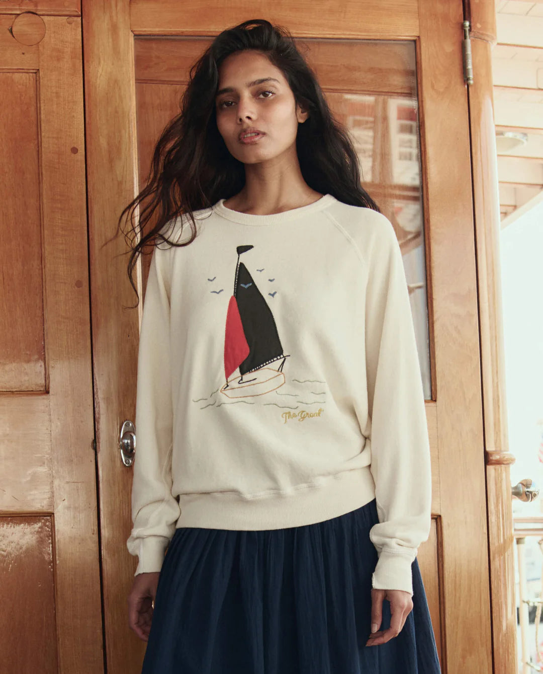 Woman wearing a cream sweatshirt with a sailboat design in front of a wooden door.