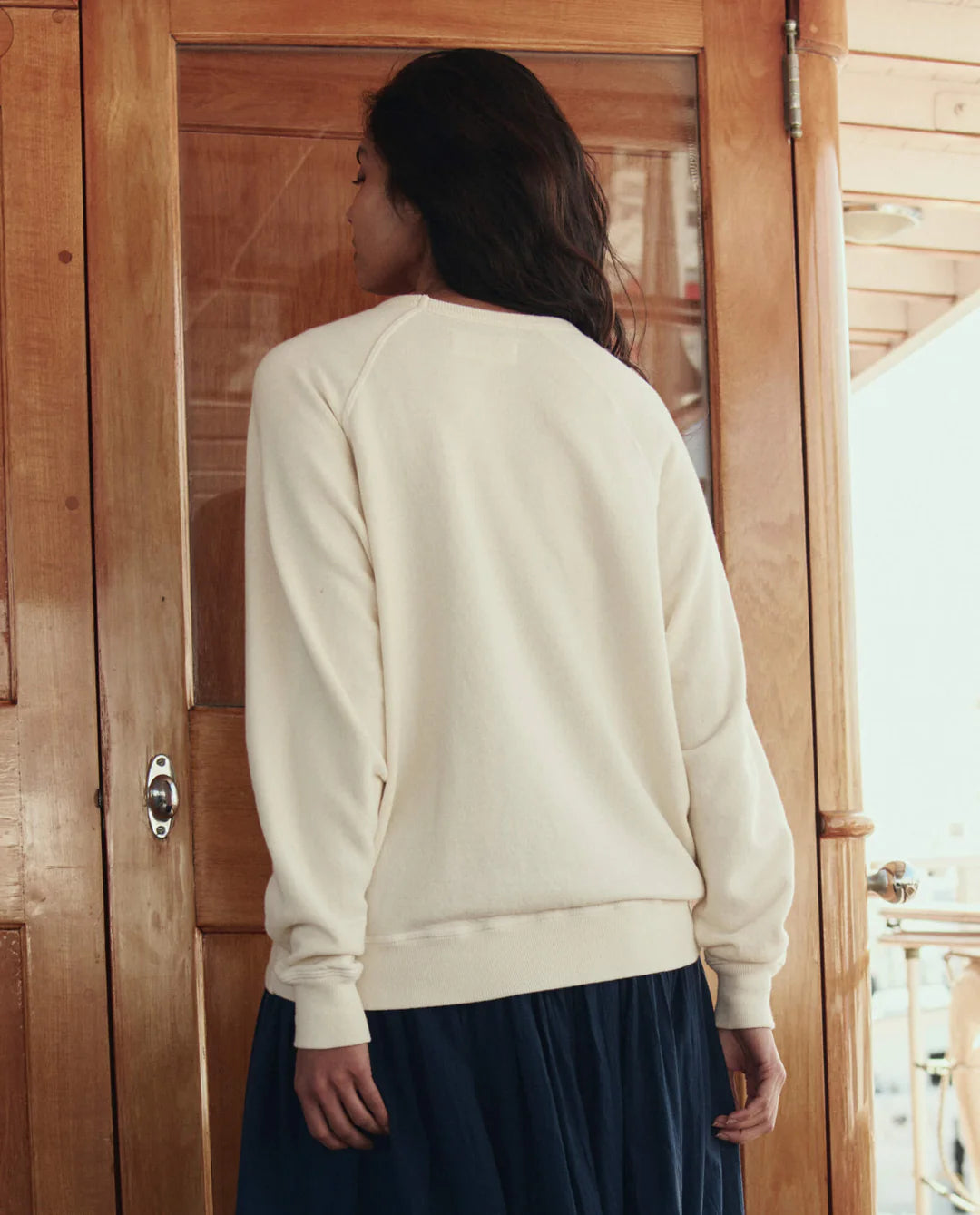 Person wearing a cream sweater and dark skirt standing in front of a wooden door.