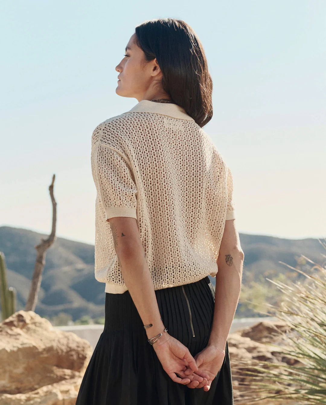 Woman wearing a beige knitted cardigan and black skirt standing in a scenic outdoor setting.