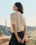 Woman wearing a beige knitted cardigan and black skirt standing in a scenic outdoor setting.