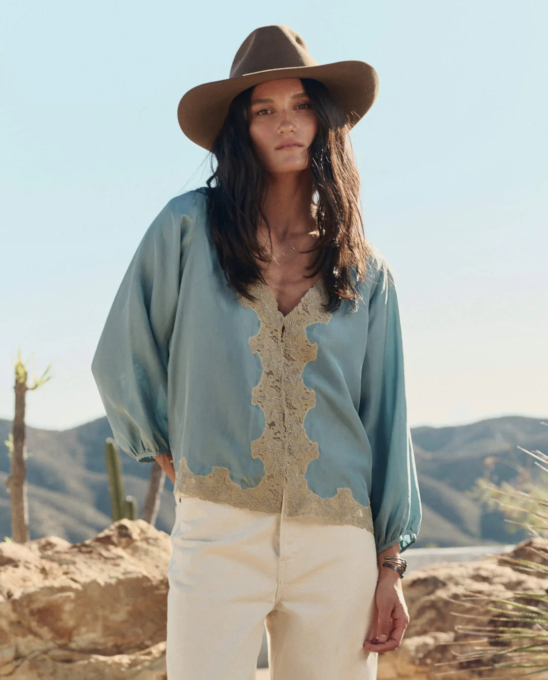 Woman wearing a light blue blouse with lace details and a wide-brimmed hat in a desert setting.