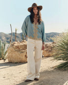 Woman in a light blue top and beige pants standing in a desert landscape with mountains in the background.