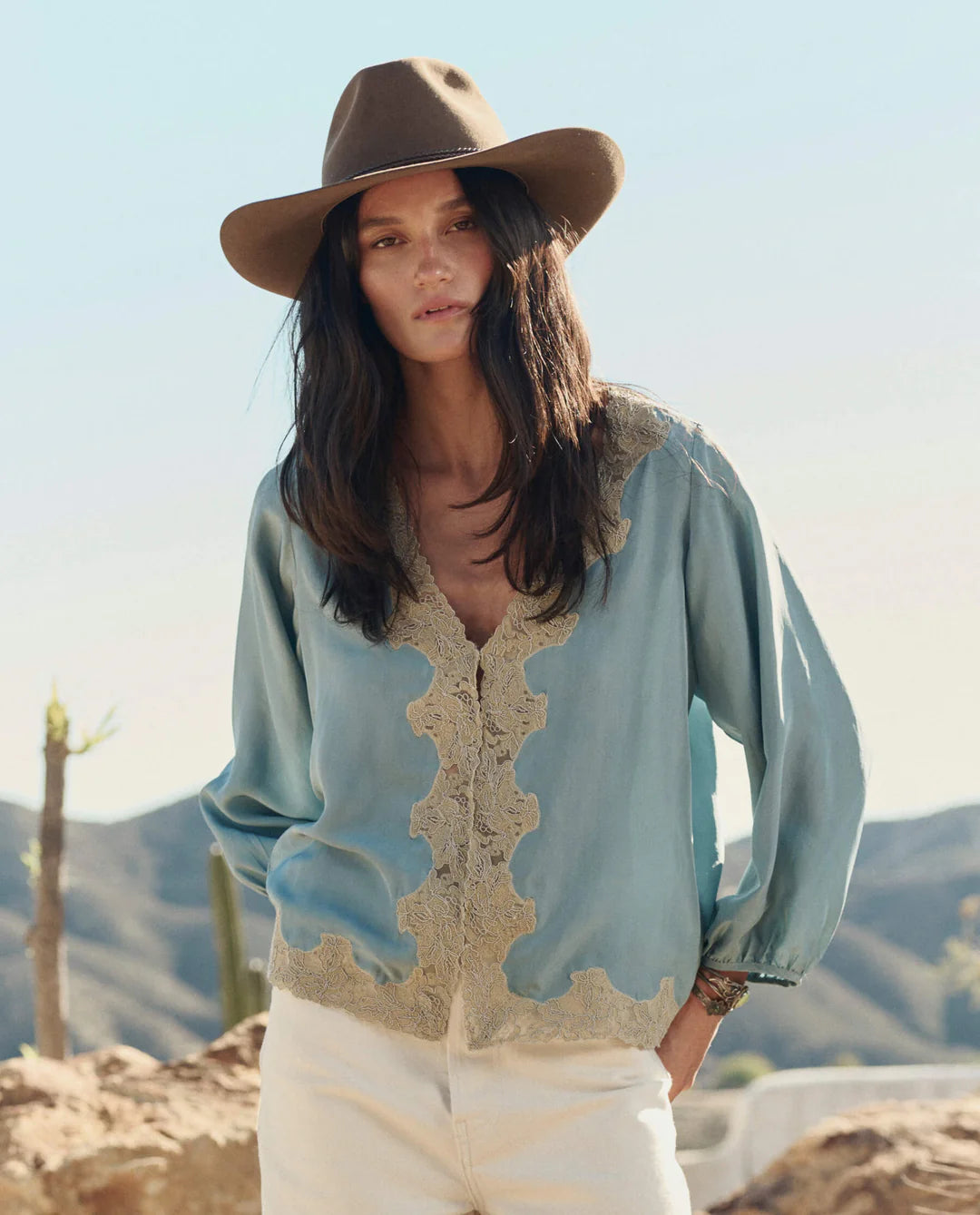 Woman wearing a light blue blouse with lace details and a brown hat in a desert setting.