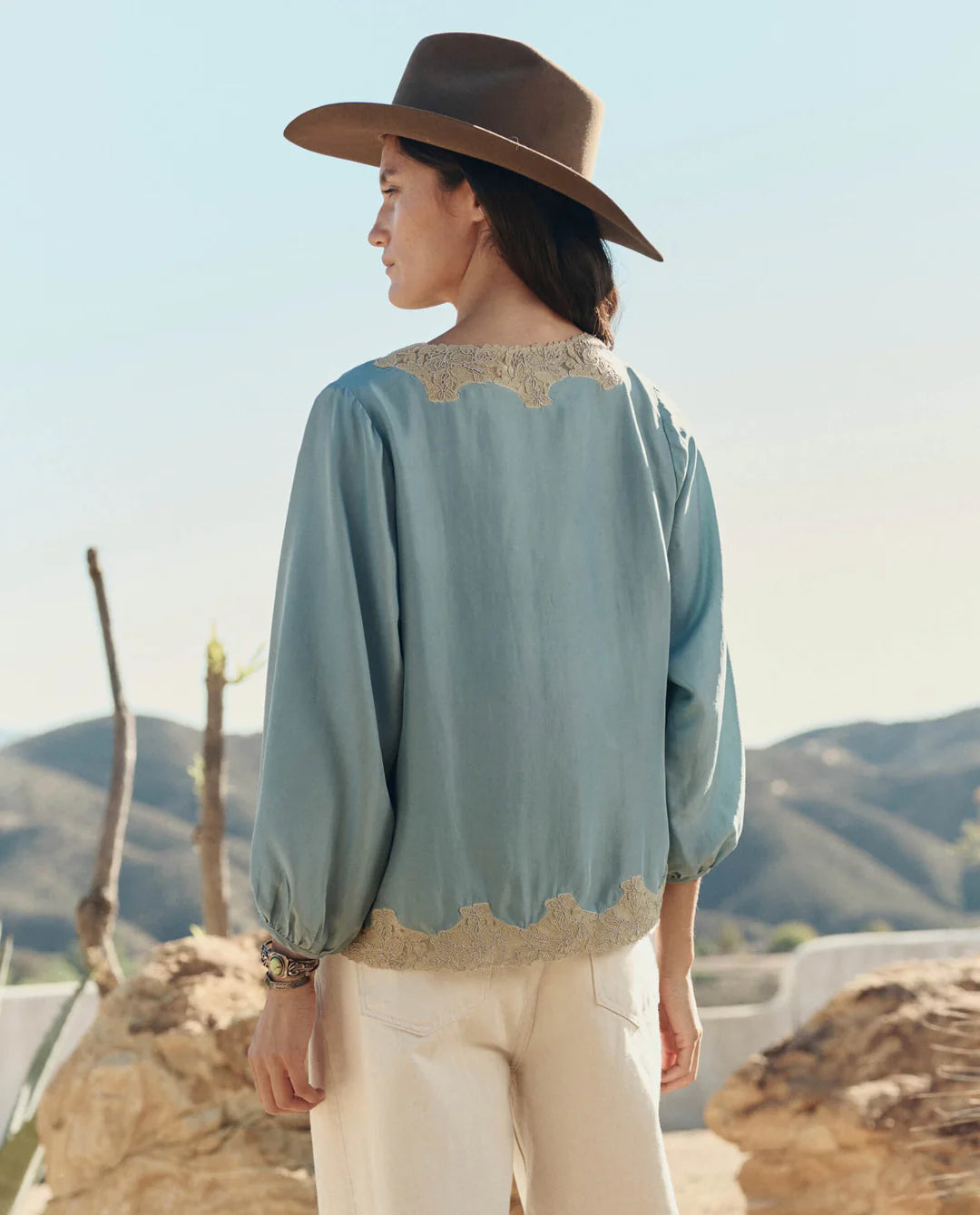 Woman wearing a light blue blouse with lace details and a brown hat, standing against a mountainous landscape.