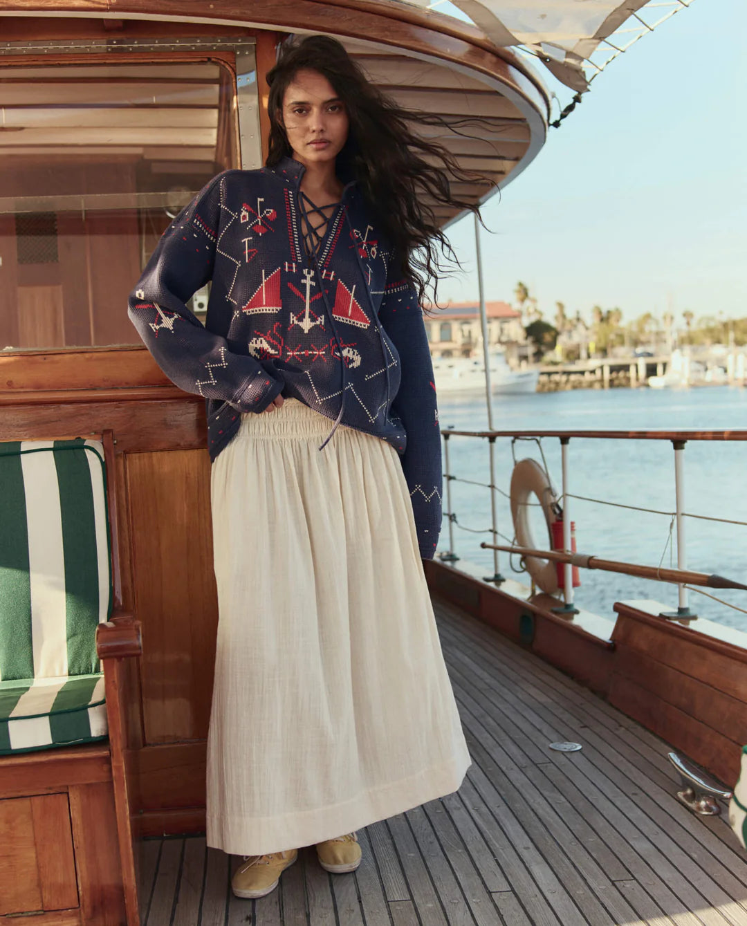 Woman on a boat deck wearing a patterned sweater and long skirt with a waterfront background