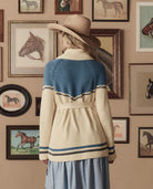 Woman wearing a patterned cardigan in front of framed horse pictures on a wall.