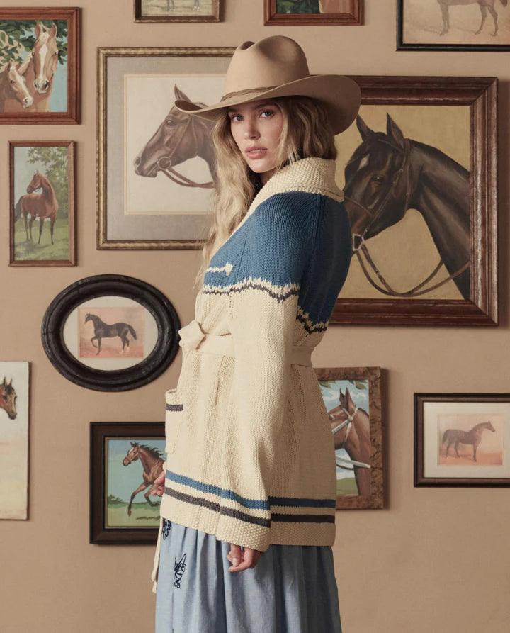 Woman wearing a patterned cardigan and hat in front of framed horse pictures on a wall.